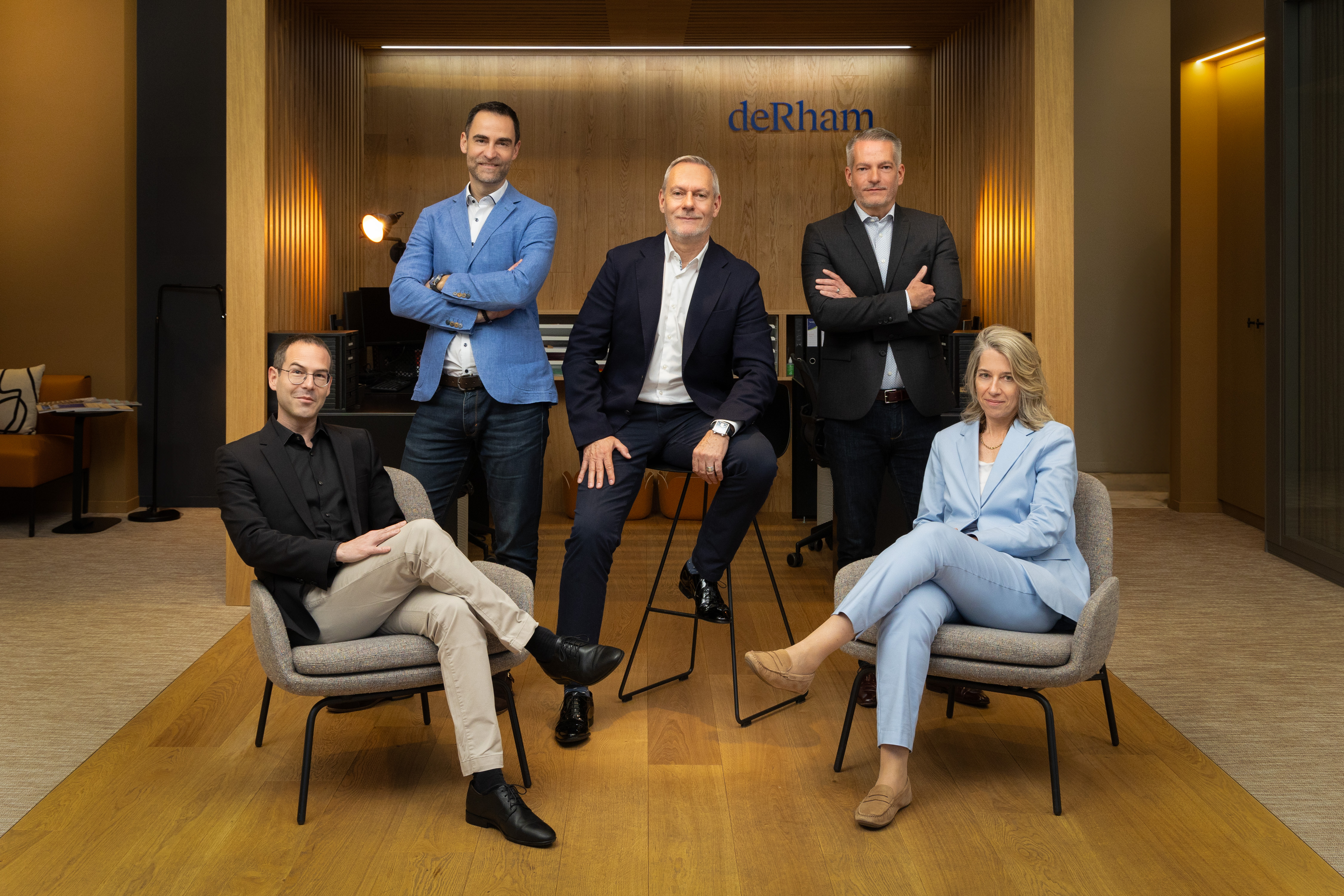 Photo de groupe montrant cinq membres de la direction de l'entreprise deRham, assis et debout dans un environnement professionnel. Deux hommes sont assis sur des fauteuils au premier plan, tandis que trois autres, dont une femme, sont debout derri&egrave;re eux. Le logo deRham est visible en arri&egrave;re-plan. &copy; deRham/Thomas Masotti